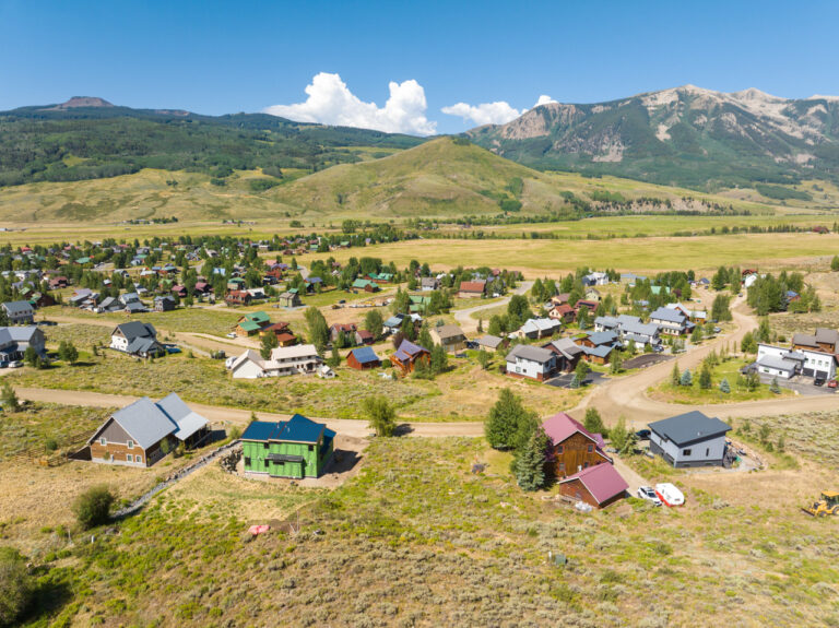 Aerial view of a small mountain town with colorful houses, dirt roads, and green fields, set against distant hills and rugged mountains.
