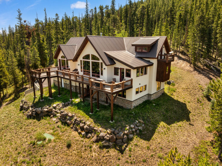 Contemporary two-story house with large wraparound deck perched on a rocky slope, surrounded by pine forest.