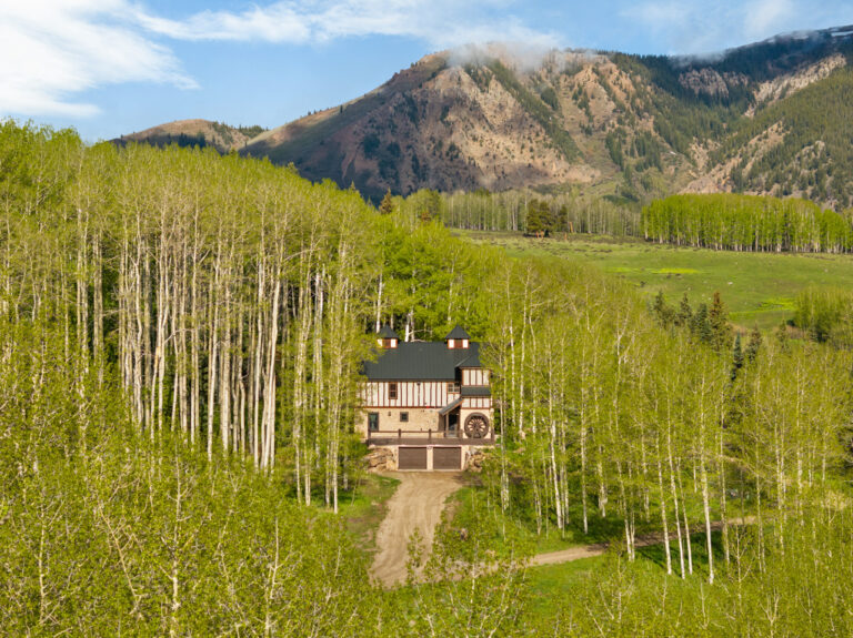 A two-story house with a dark green roof nestled among tall aspen trees, mountain backdrop in a valley.