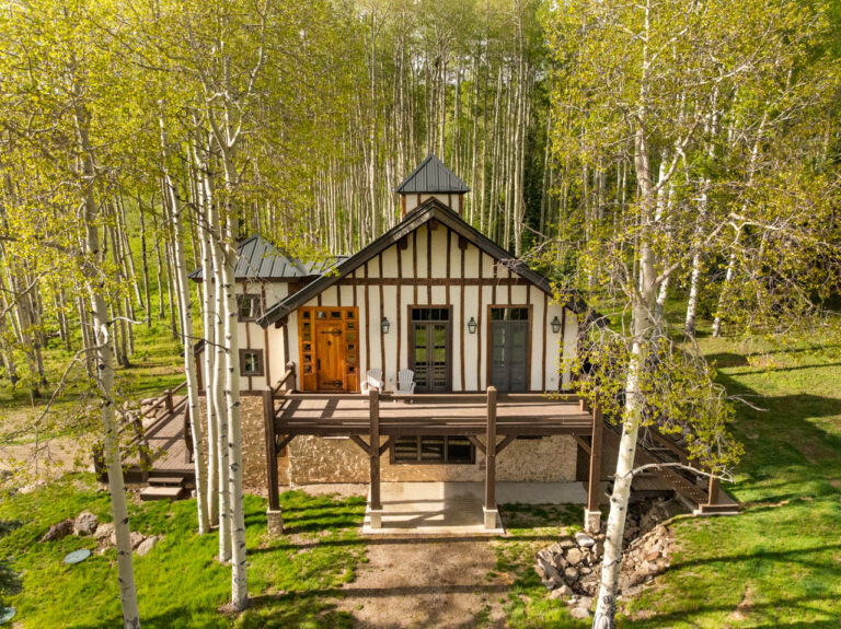 A Tudor-style house with white walls and dark wooden beams on a raised deck among aspen trees in a forest setting (a charming cabin).