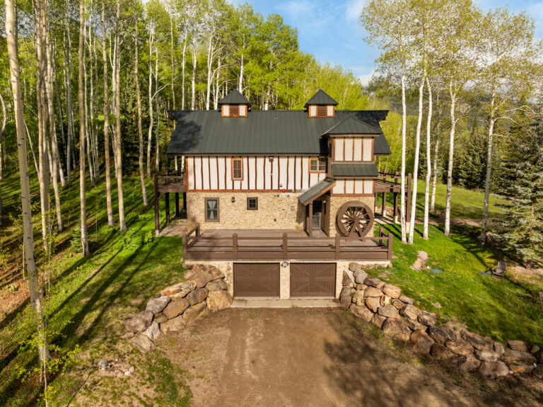 Rustic timber-and-stone house in a forest setting with a water wheel and a two-car garage below.