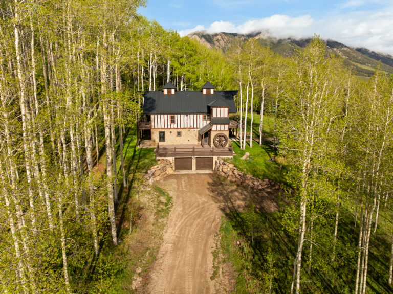 A rustic two-story house with a dark roof and stone foundation, a wooden deck and a waterwheel on the side, set in a dense aspen forest with a dirt driveway leading up to it.