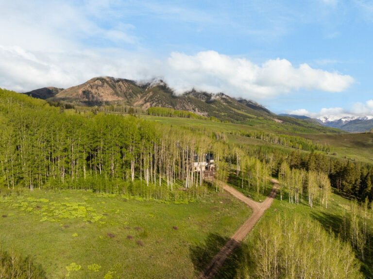 A rural valley with a dirt road winding toward a house nestled among an aspen grove, with green hills and a mountain range under a blue sky.