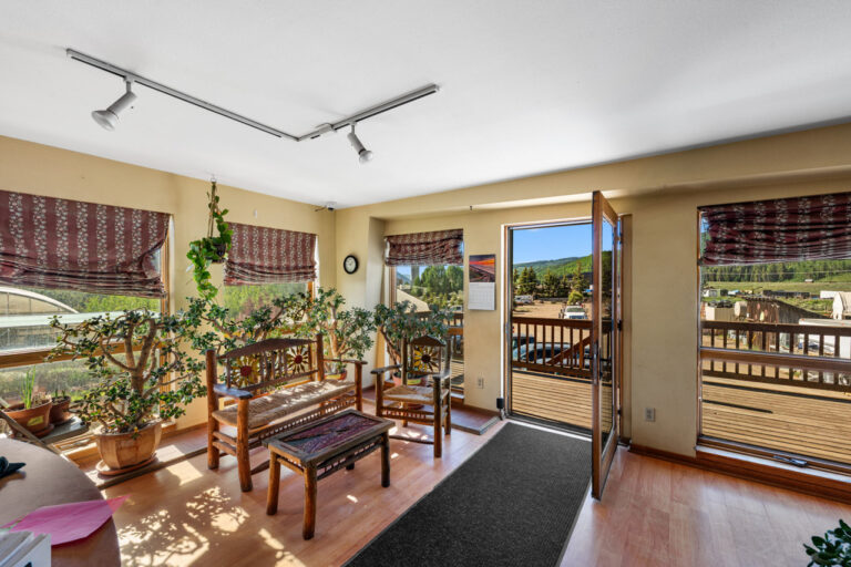 Sunlit sitting area with wooden chairs, a coffee table, and potted plants near large windows overlooking a deck and mountains beyond