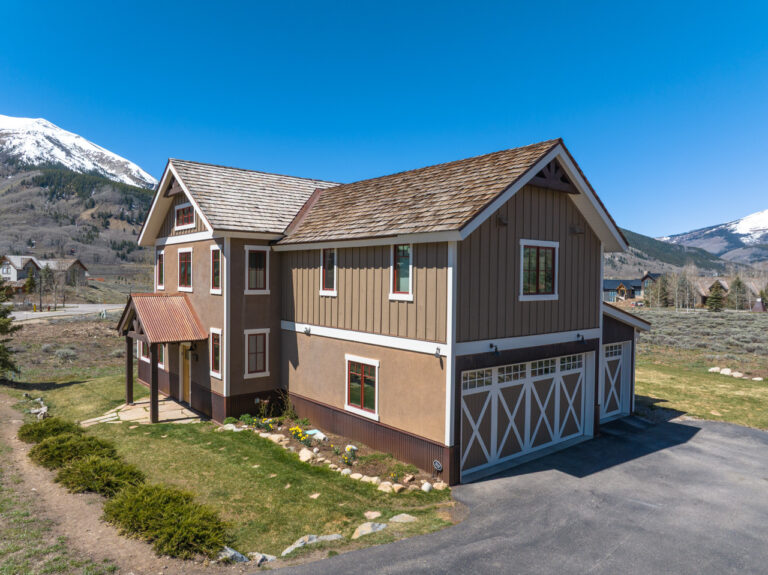 Two-story house with a three-car garage in a mountain landscape under a bright blue sky.