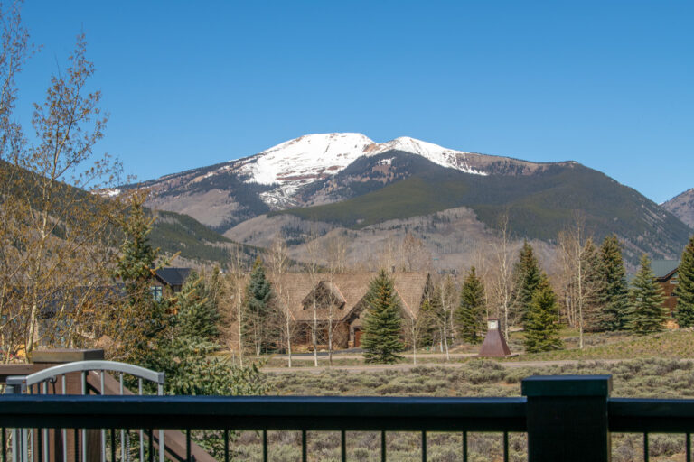 Snowy mountain peak under a clear blue sky, with evergreens and chalet-style houses in a valley viewed from a railinged deck.