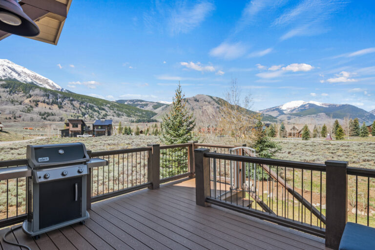 Deck with a gas grill on the left, railing in the foreground, and snow-capped mountains under a blue sky.