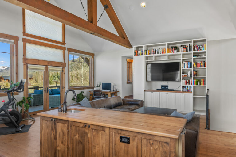 Open-concept living room with exposed wooden beams, large windows, and a built-in bookcase around a wall-mounted TV.
