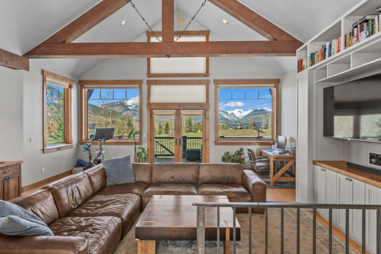Cozy living room with a brown leather sectional, wooden coffee table, and large windows showing snowy mountain views.