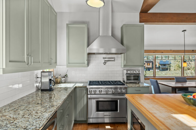 Open-concept kitchen with sage green cabinets, stainless steel gas range and vent hood, white subway tile backsplash, and granite countertops.