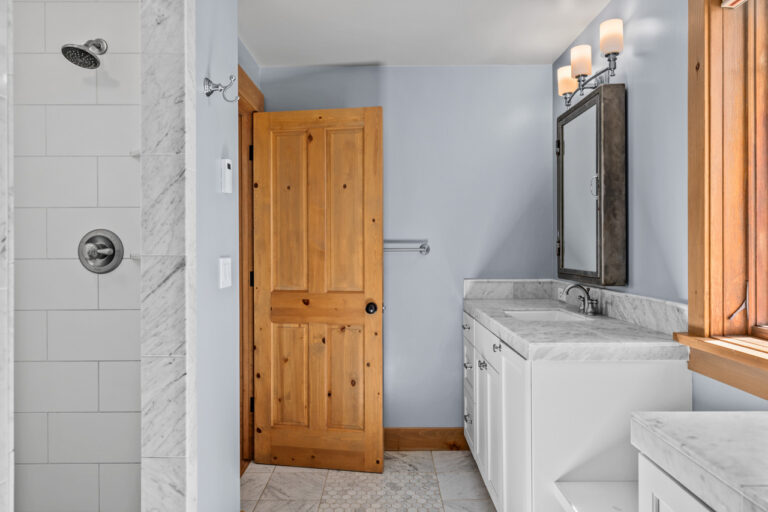 Bathroom with light blue walls, white marble countertop, and a wooden door; vanity with sink, framed mirror, and three-light fixture.