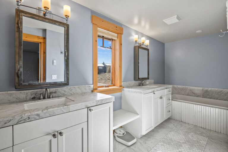 Bathroom with two white vanities, marble countertops, and a large window between sinks; blue-gray walls and marble floor tiles reflect natural light.