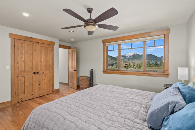 Bedroom with a large wooden-framed window showing mountains, a gray quilted bed, and a ceiling fan above.