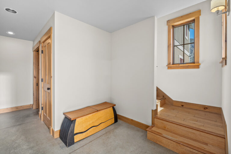 Bright entryway with white walls, wood trim, a curved storage bench, and a wooden staircase nearby. A window shows the mountain exterior.