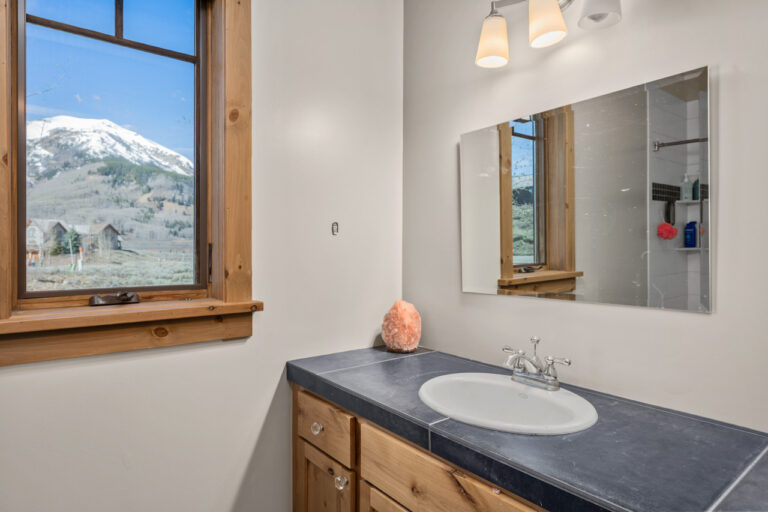 Bathroom vanity with dark countertop, white oval sink, chrome faucet, wooden cabinets; salt lamp on counter; mountain-view window.