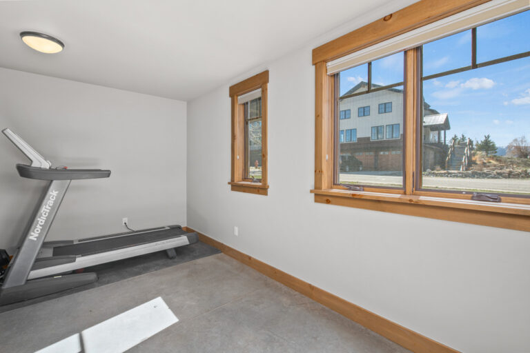A small home gym with a NordickTrack treadmill against a gray wall, and two large windows with wooden frames letting in daylight.