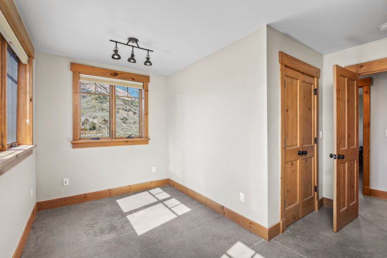 Sunlit empty bedroom with grey concrete-like floor, natural wood trim, and a large window; two wooden doors on the right.