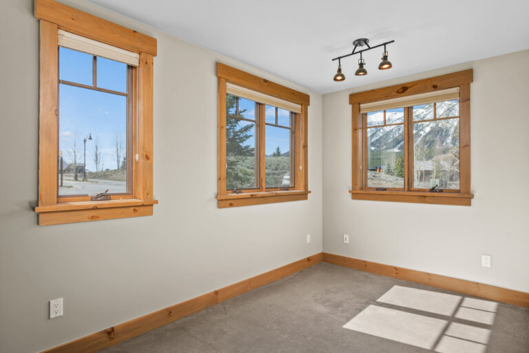 Empty room with three wooden-framed windows, beige walls, and baseboards; sunlight streams in.