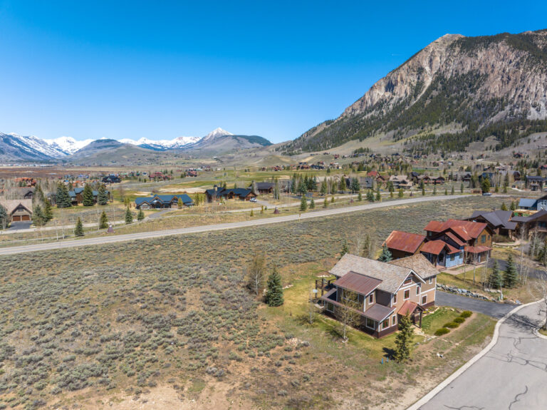 A mountain valley town with houses along a curved road and a large hillside, snow-capped peaks in the distance under a clear blue sky.