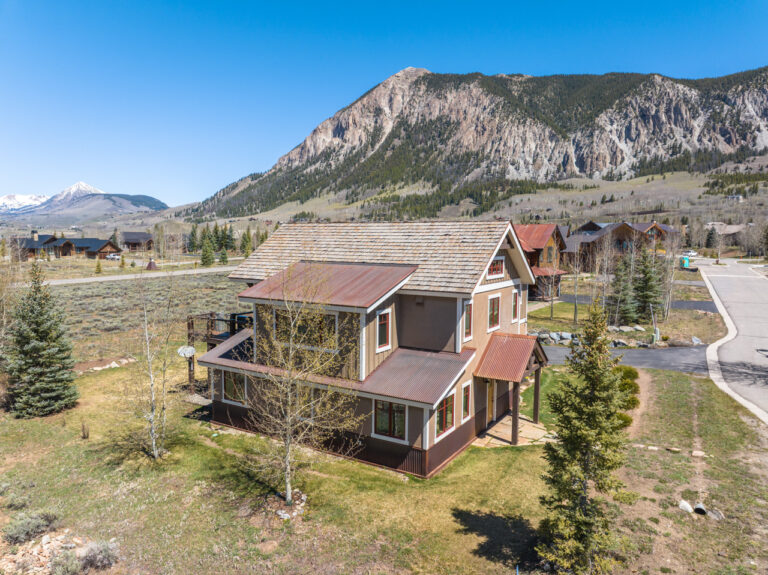Two-story alpine home with wood siding and a metal roof in a mountain neighborhood, with rocky cliffs in the background.