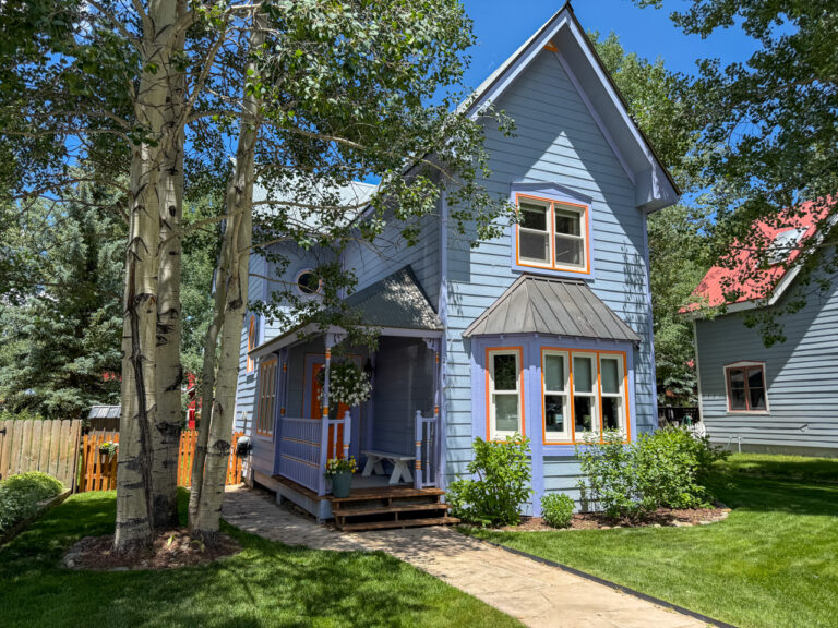 Blue two-story house with orange trim and a small covered porch, front yard with green lawn and trees.