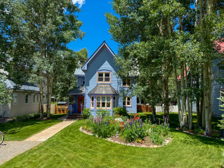 Blue two-story cottage with orange-trimmed windows, a glass bay window, and a stone path leading to the front door, set among a colorful flower bed and tall trees.