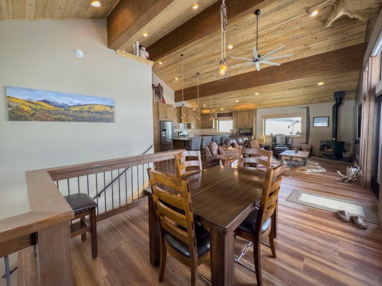 Open-concept cabin interior with a dark wood dining table and six chairs overlooking a living area with a fireplace and vaulted wooden ceiling.