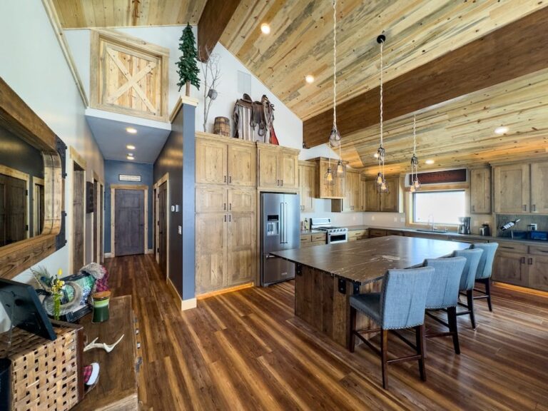 Rustic kitchen with a large wooden island and four blue upholstered stools, stainless appliances, and exposed wood beam ceiling.