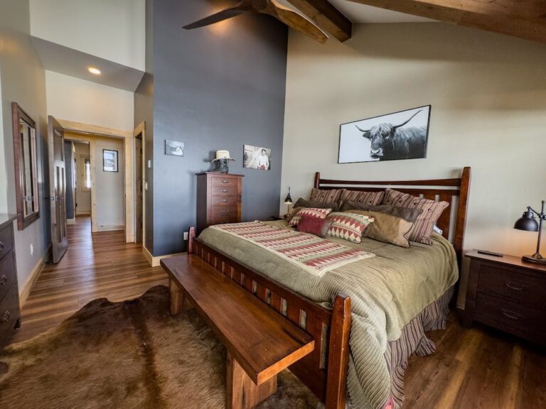 Rustic bedroom with a large wooden bed, matching dressers, bench at the foot, cow artwork, and warm wood tones.