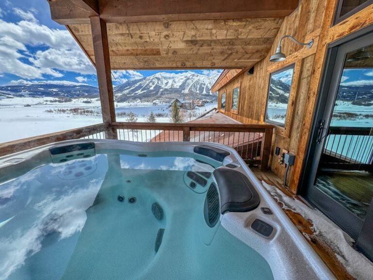Outdoor hot tub on a wooden deck with a snowy mountain and lake view under a blue sky.