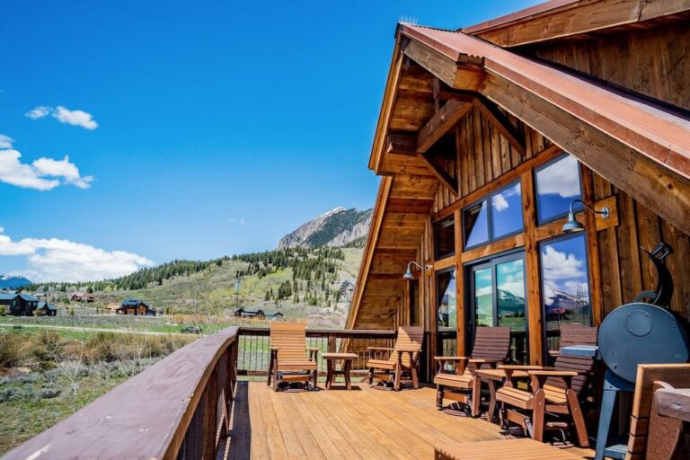 Wooden cabin with large glass windows and an outdoor deck, overlooking green hills and distant mountains.