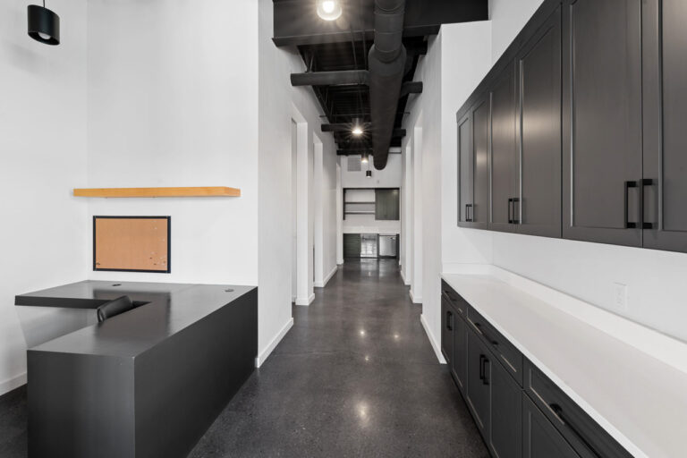 Long industrial-style office hallway with black cabinetry along the right and a dark reception desk at the left; exposed ducts and pendant lights above.