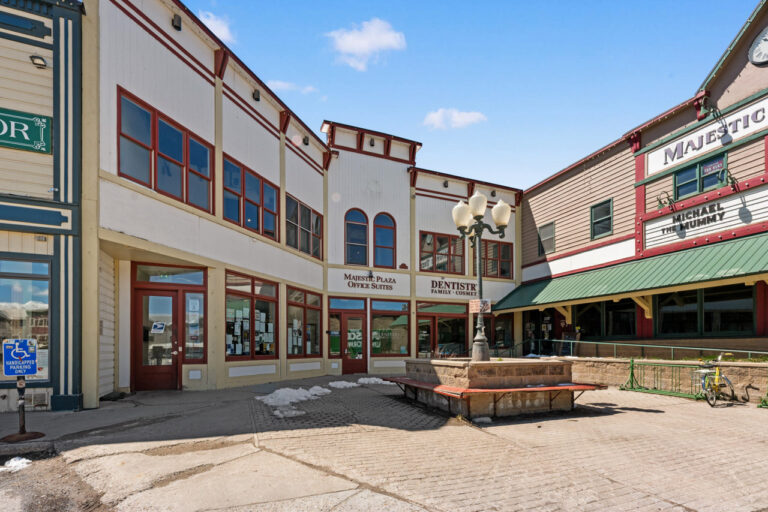 Small-town plaza with white buildings trimmed in red, a central stone planter and vintage lamppost; storefronts read Majestic Plaza Office Suites and Dentistry.