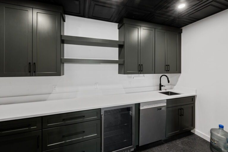 Dark kitchen with green-gray cabinets, white countertop, and a black faucet over a stainless sink, plus a dishwasher and wine cooler under the counter.