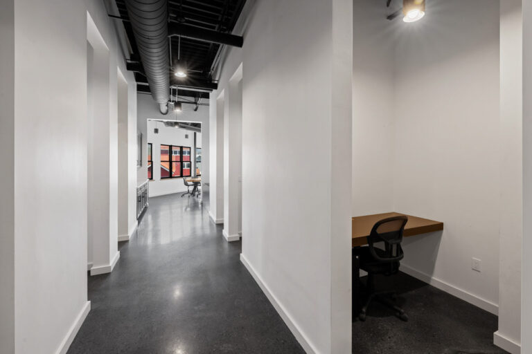 Long rectangular office hallway with white walls, exposed black ceiling ducts, and a desk alcove on the right.