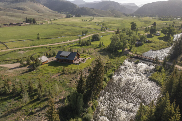 Aerial view of a rural valley with green fields, a winding river, and a wooden bridge near scattered houses and distant hills and mountains.
