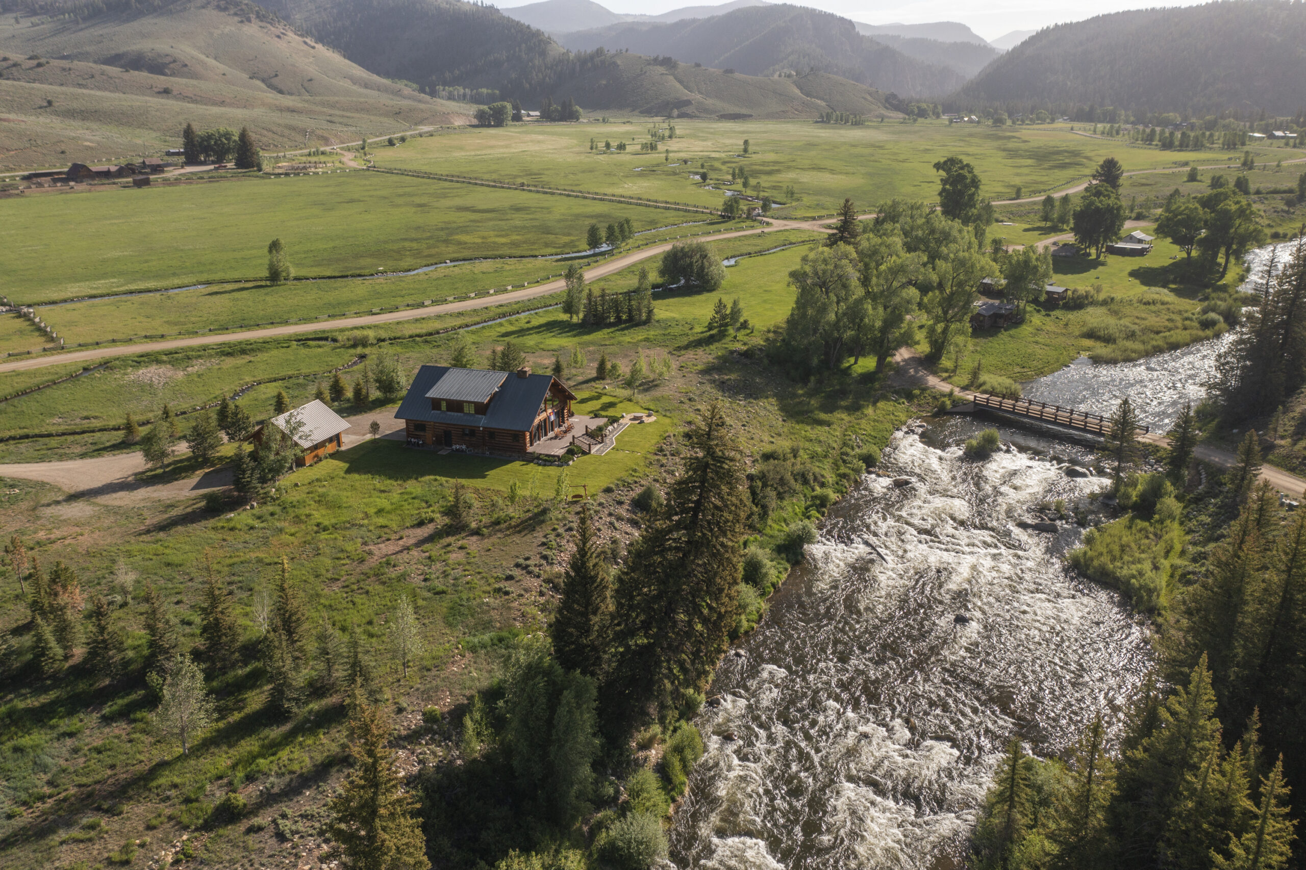 Aerial view of a rural valley with green fields, a winding river, and a wooden bridge near scattered houses and distant hills and mountains.
