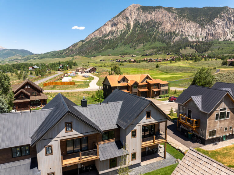 Aerial view of rustic mountain cabins in a valley, with a towering rocky peak and green slopes behind them.